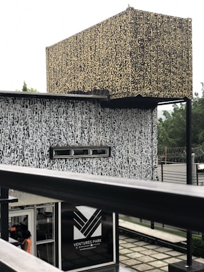 An architectural structure with complex, abstract patterns covering its surfaces. The design is characterized by a combination of beige and black colors. It has a rectangular sign labeled 'Ventures Park' near the bottom with an entrance visible just below, where a person is partially visible. The background features some greenery with a slightly blurred appearance.