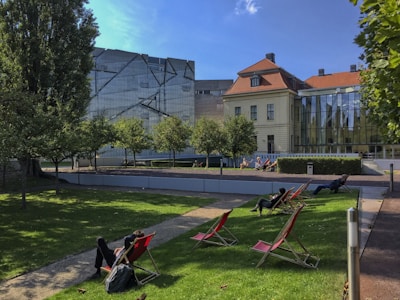 A modern building with a geometric design stands adjacent to a classic brick structure with a red roof. Trees and a well-maintained lawn with scattered deck chairs are in the foreground, where a few people relax under the blue sky.