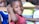 A young child wearing a red shirt smiles warmly while resting their arm on the back of a wooden chair. Another child in a blue shirt is partially visible in the background, along with blurred figures in various colorful clothing.