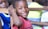 A young child wearing a red shirt smiles warmly while resting their arm on the back of a wooden chair. Another child in a blue shirt is partially visible in the background, along with blurred figures in various colorful clothing.