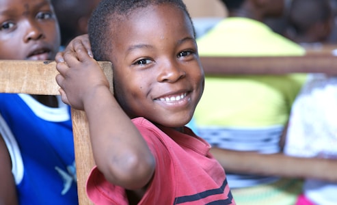 A young child wearing a red shirt smiles warmly while resting their arm on the back of a wooden chair. Another child in a blue shirt is partially visible in the background, along with blurred figures in various colorful clothing.