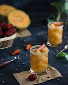 Two glasses filled with yellow melon balls and garnished with strawberry slices sit on a dark surface, surrounded by scattered strawberries, whole melons, and kitchen utensils. The glass rims are coated with sugar, adding a touch of elegance to the presentation.