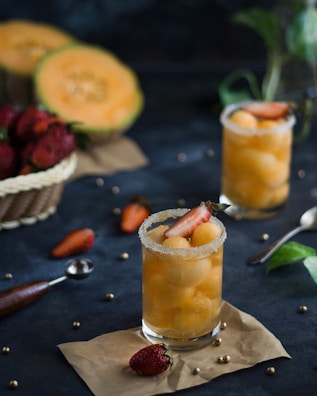 Two glasses filled with yellow melon balls and garnished with strawberry slices sit on a dark surface, surrounded by scattered strawberries, whole melons, and kitchen utensils. The glass rims are coated with sugar, adding a touch of elegance to the presentation.
