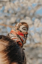 A close-up of a patterned scarf tied around a section of hair. The scarf features intricate designs in shades of orange, brown, black, and white, creating a stylish accessory. The background is a blurred out-of-focus view of light-colored blossoms against a blue sky.