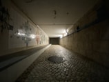 A dimly lit tunnel with cobblestone flooring and textured walls. The left wall features white tiles with graffiti and mixed colors, while the right wall has exposed brickwork. A circular metal grate is embedded in the floor, and blue pipes run along the ceiling.