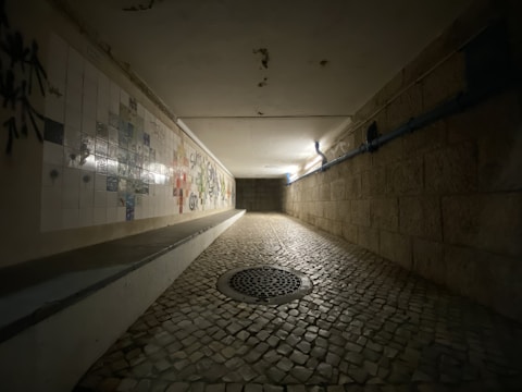 A dimly lit tunnel with cobblestone flooring and textured walls. The left wall features white tiles with graffiti and mixed colors, while the right wall has exposed brickwork. A circular metal grate is embedded in the floor, and blue pipes run along the ceiling.