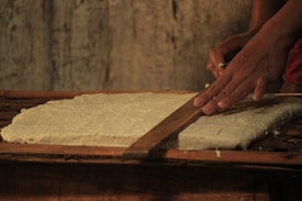 Hands are seen carefully slicing a large slab of dough-like substance with a wooden tool, against a rustic wooden background.