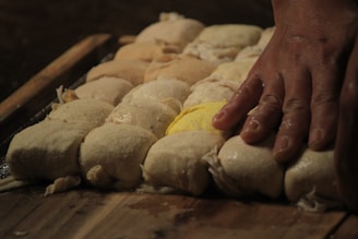 A hand pressing down on a batch of dough balls on a wooden surface, with one of the dough balls having a yellow tint.