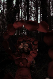 A woman is holding a string of red rose petals while standing in a shadowy forest. Her expression is contemplative, and the petals form a loose oval shape around her. The trees in the background are tall and dense, casting dark shadows.