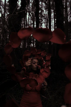 A woman is holding a string of red rose petals while standing in a shadowy forest. Her expression is contemplative, and the petals form a loose oval shape around her. The trees in the background are tall and dense, casting dark shadows.