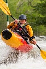 A kayaker navigates through white water rapids, gripping a paddle with both hands. The person is wearing a helmet and a yellow and red kayak jacket. Water splashes around them, creating a dynamic and intense scene. Surrounding them is a backdrop of lush, green foliage suggesting a natural, forested river setting.
