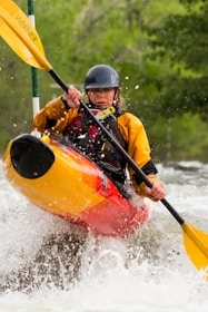 A kayaker navigates through white water rapids, gripping a paddle with both hands. The person is wearing a helmet and a yellow and red kayak jacket. Water splashes around them, creating a dynamic and intense scene. Surrounding them is a backdrop of lush, green foliage suggesting a natural, forested river setting.