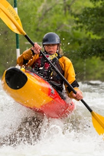 A kayaker navigates through white water rapids, gripping a paddle with both hands. The person is wearing a helmet and a yellow and red kayak jacket. Water splashes around them, creating a dynamic and intense scene. Surrounding them is a backdrop of lush, green foliage suggesting a natural, forested river setting.