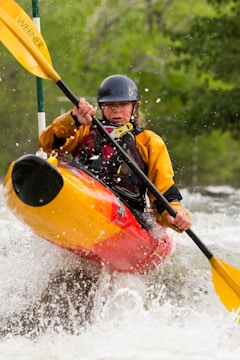 A kayaker navigates through white water rapids, gripping a paddle with both hands. The person is wearing a helmet and a yellow and red kayak jacket. Water splashes around them, creating a dynamic and intense scene. Surrounding them is a backdrop of lush, green foliage suggesting a natural, forested river setting.
