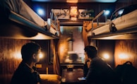 A cozy train cabin with two bunk beds and dim lighting, featuring two people seated on opposite beds, gazing out of the window. The interior is warmly lit, with wooden furnishings and soft, yellow lamps. Pictures hang on the wall above the window, adding to the intimate atmosphere.