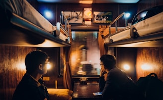 A cozy train cabin with two bunk beds and dim lighting, featuring two people seated on opposite beds, gazing out of the window. The interior is warmly lit, with wooden furnishings and soft, yellow lamps. Pictures hang on the wall above the window, adding to the intimate atmosphere.