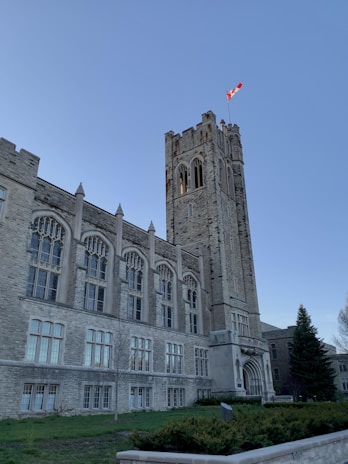 A large, historical stone building with Gothic architecture is visible. It features tall arched windows and a prominent tower with intricate stonework. The Canadian flag is flying atop the tower. There is a neatly trimmed green lawn in the foreground and a tall evergreen tree on the right.