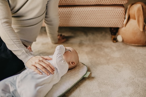 A baby lies on a cushioned mat on the floor, partially shaded by an arm from an adult wearing a sweater. The setting includes a patterned sofa and a plush toy.