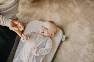 A baby lying on a cushioned mat on a carpeted floor, dressed in a white onesie and holding an adult's finger. The baby has a pacifier in its mouth and is gazing upwards.