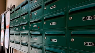 A row of green metal mailboxes with numbered labels are mounted on a wall. Each mailbox has a lock and a small handle for opening. The numbers range from 201 to 402. The boxes are positioned next to some posters attached to a grid-like structure on the left edge of the image.