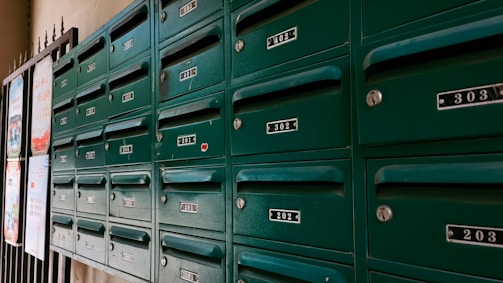 A row of green metal mailboxes with numbered labels are mounted on a wall. Each mailbox has a lock and a small handle for opening. The numbers range from 201 to 402. The boxes are positioned next to some posters attached to a grid-like structure on the left edge of the image.