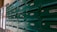 A row of green metal mailboxes with numbered labels are mounted on a wall. Each mailbox has a lock and a small handle for opening. The numbers range from 201 to 402. The boxes are positioned next to some posters attached to a grid-like structure on the left edge of the image.