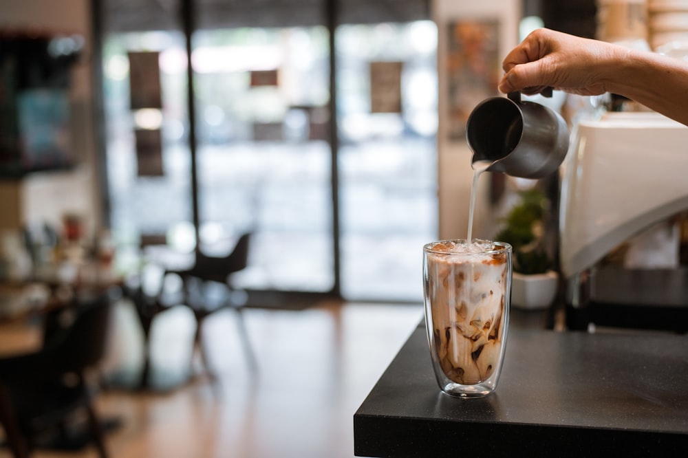 person pouring brown liquid on clear drinking glass