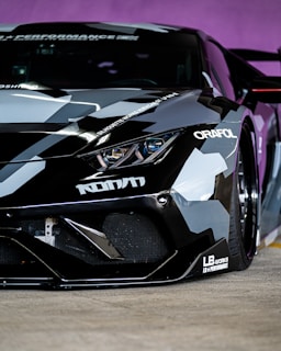A close-up of the front end of a sleek, modified sports car featuring a custom paint job with geometric camouflage patterns in shades of gray, black, and white. The car is adorned with various decals and branding, including 'ORAFL' and 'LB WORKS'. The angular headlights and aerodynamic design emphasize its high-performance nature.