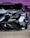 A close-up of the front end of a sleek, modified sports car featuring a custom paint job with geometric camouflage patterns in shades of gray, black, and white. The car is adorned with various decals and branding, including 'ORAFL' and 'LB WORKS'. The angular headlights and aerodynamic design emphasize its high-performance nature.