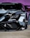 A close-up of the front end of a sleek, modified sports car featuring a custom paint job with geometric camouflage patterns in shades of gray, black, and white. The car is adorned with various decals and branding, including 'ORAFL' and 'LB WORKS'. The angular headlights and aerodynamic design emphasize its high-performance nature.