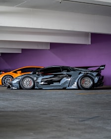 A sleek, customized black sports car with a wide body kit and aggressive styling is parked in a garage. The background features a vibrant purple wall and another orange sports car is partially visible behind the black car.