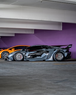 A sleek, customized black sports car with a wide body kit and aggressive styling is parked in a garage. The background features a vibrant purple wall and another orange sports car is partially visible behind the black car.