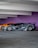 A sleek, customized black sports car with a wide body kit and aggressive styling is parked in a garage. The background features a vibrant purple wall and another orange sports car is partially visible behind the black car.