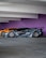 A sleek, customized black sports car with a wide body kit and aggressive styling is parked in a garage. The background features a vibrant purple wall and another orange sports car is partially visible behind the black car.