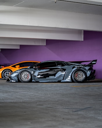 A sleek, customized black sports car with a wide body kit and aggressive styling is parked in a garage. The background features a vibrant purple wall and another orange sports car is partially visible behind the black car.