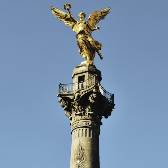 A tall, ornate column topped with a golden winged statue stands prominently against a clear blue sky. The statue is poised holding a laurel wreath.