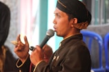 A man wearing traditional clothing, including a dark-colored outfit with decorative buttons and a headscarf, is speaking into a microphone. He is visible from the side and is gesturing with his hand while holding an object resembling a ring or seal. There are blurred background elements, including blue plastic chairs and a mix of light and dark hues.