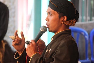 A man wearing traditional clothing, including a dark-colored outfit with decorative buttons and a headscarf, is speaking into a microphone. He is visible from the side and is gesturing with his hand while holding an object resembling a ring or seal. There are blurred background elements, including blue plastic chairs and a mix of light and dark hues.