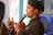A man wearing traditional clothing, including a dark-colored outfit with decorative buttons and a headscarf, is speaking into a microphone. He is visible from the side and is gesturing with his hand while holding an object resembling a ring or seal. There are blurred background elements, including blue plastic chairs and a mix of light and dark hues.