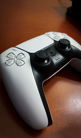 A gaming controller is placed on a wooden surface, featuring a sleek black and white design with prominent joysticks and buttons.