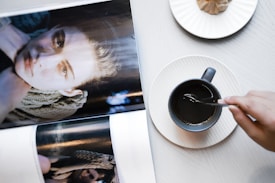 A table displays an open magazine featuring a close-up of a person's face, with space between them showing more of the magazine. Beside the magazine is a saucer and a dark mug filled with a beverage being stirred by a hand. A plate with a pastry or snack is also visible.