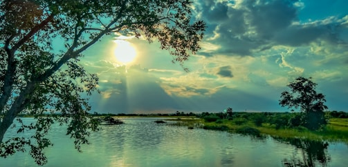 The image features a serene landscape with a large body of water in the foreground, reflecting the vibrant colors of the sky. Sunlight breaks through the clouds, creating a dramatic effect and illuminating the scene. Tall trees frame the image on both sides, adding depth and natural beauty.