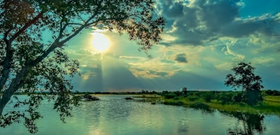 The image features a serene landscape with a large body of water in the foreground, reflecting the vibrant colors of the sky. Sunlight breaks through the clouds, creating a dramatic effect and illuminating the scene. Tall trees frame the image on both sides, adding depth and natural beauty.