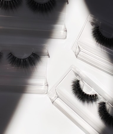 False eyelashes arranged in individual transparent plastic cases lying on a white surface with shadows and light creating a contrast.