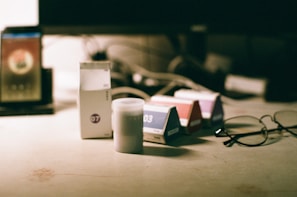 Various boxes and a small cylindrical container are placed on a light-colored surface. To the right, a pair of eyeglasses is resting on the surface. In the background, a blurred monitor screen and additional objects are visible.