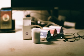 Various boxes and a small cylindrical container are placed on a light-colored surface. To the right, a pair of eyeglasses is resting on the surface. In the background, a blurred monitor screen and additional objects are visible.