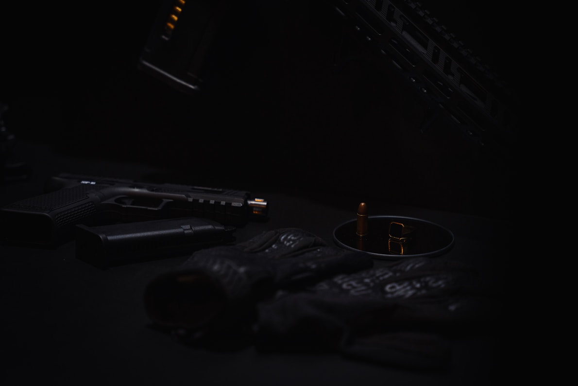 A dark and moody composition features a handgun with a silencer, ammunition, and tactical gloves organized on a surface. The subdued lighting casts shadows, creating a mysterious atmosphere.