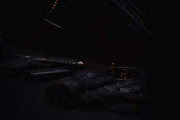 A dark and moody composition features a handgun with a silencer, ammunition, and tactical gloves organized on a surface. The subdued lighting casts shadows, creating a mysterious atmosphere.