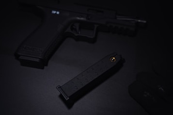 A dark image featuring a handgun placed on a black surface. A magazine is partially visible next to it, with a glove lying nearby. The scene is dimly lit, creating an atmosphere of seriousness and tension.