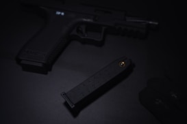 A dark image featuring a handgun placed on a black surface. A magazine is partially visible next to it, with a glove lying nearby. The scene is dimly lit, creating an atmosphere of seriousness and tension.